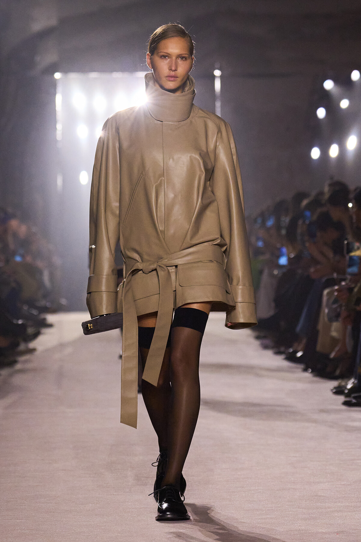 Model on a fashion runway wearing a beige oversized leather coat with a wide belt, black stockings, and black shoes.