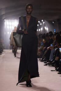 Model on the runway wearing a long dark wrap dress, carrying a large gray tote over her shoulder.