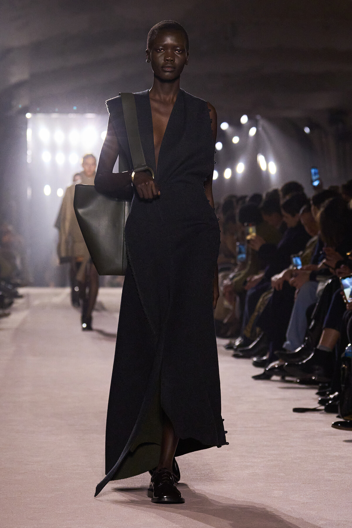 Model on the runway wearing a long dark wrap dress, carrying a large gray tote over her shoulder.