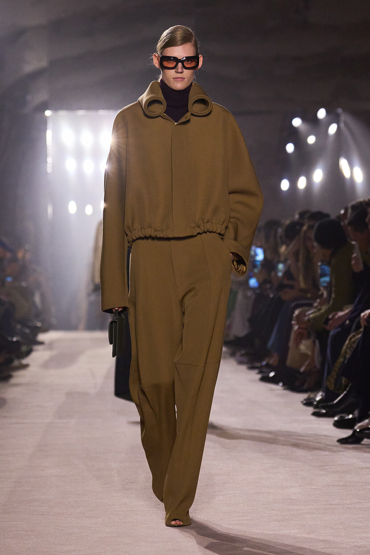 Model on a runway wearing a camel-toned coordinated set: oversized top with a cinched waist and matching wide-leg pants, black turtleneck, large sunglasses, and a small dark handbag.