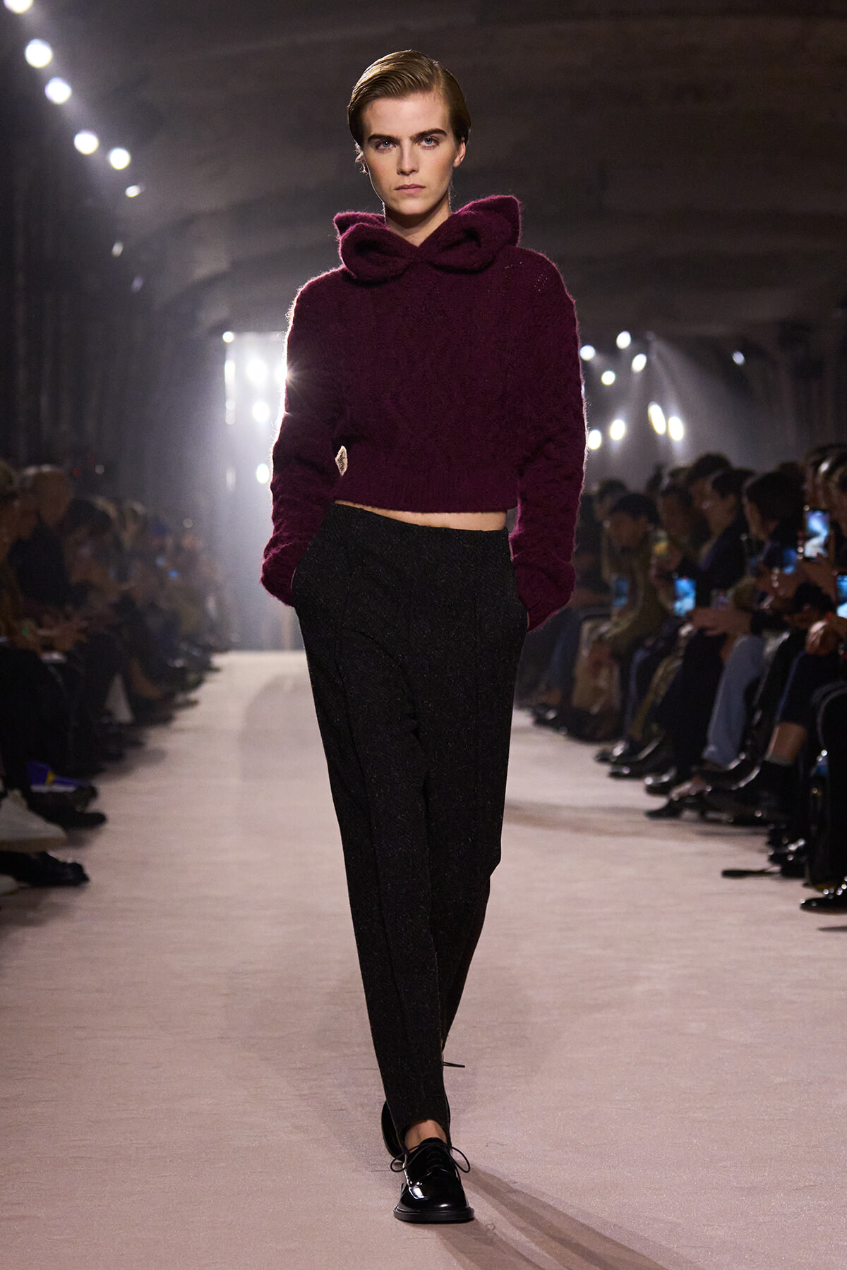Model on the runway wearing a cropped burgundy fuzzy sweater with a large hood and black tailored pants.