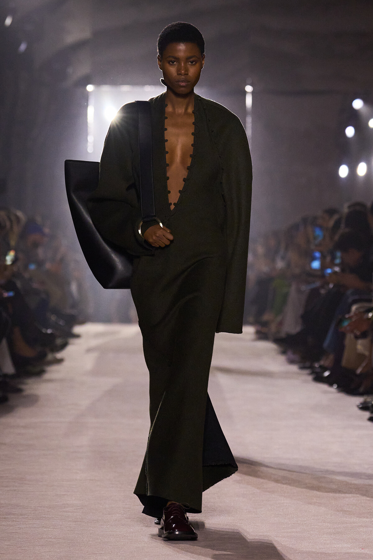 Model walks the runway in an olive-green, deep-plunge jumpsuit with a large black shoulder bag and burgundy shoes, under studio lights.