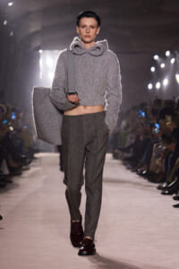 Model on a runway wearing a gray fuzzy cropped sweater with a large hood, a matching tote bag, plaid trousers, and burgundy shoes.