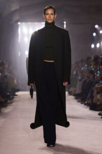 Model on a runway wearing a long black coat over a dark turtleneck and wide-leg pants, holding a small handbag.