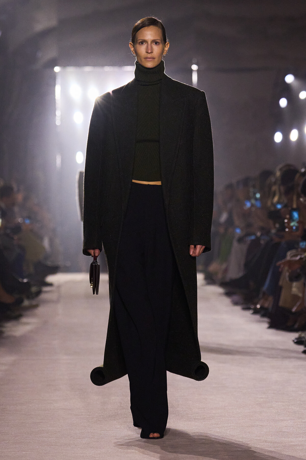 Model on a runway wearing a long black coat over a dark turtleneck and wide-leg pants, holding a small handbag.