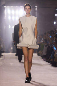 Model walks a runway in a minimalist sleeveless ivory shift dress with a high, round neckline and sculpted shoulders, paired with black lace-up shoes and a rolled fabric accessory at the hem.