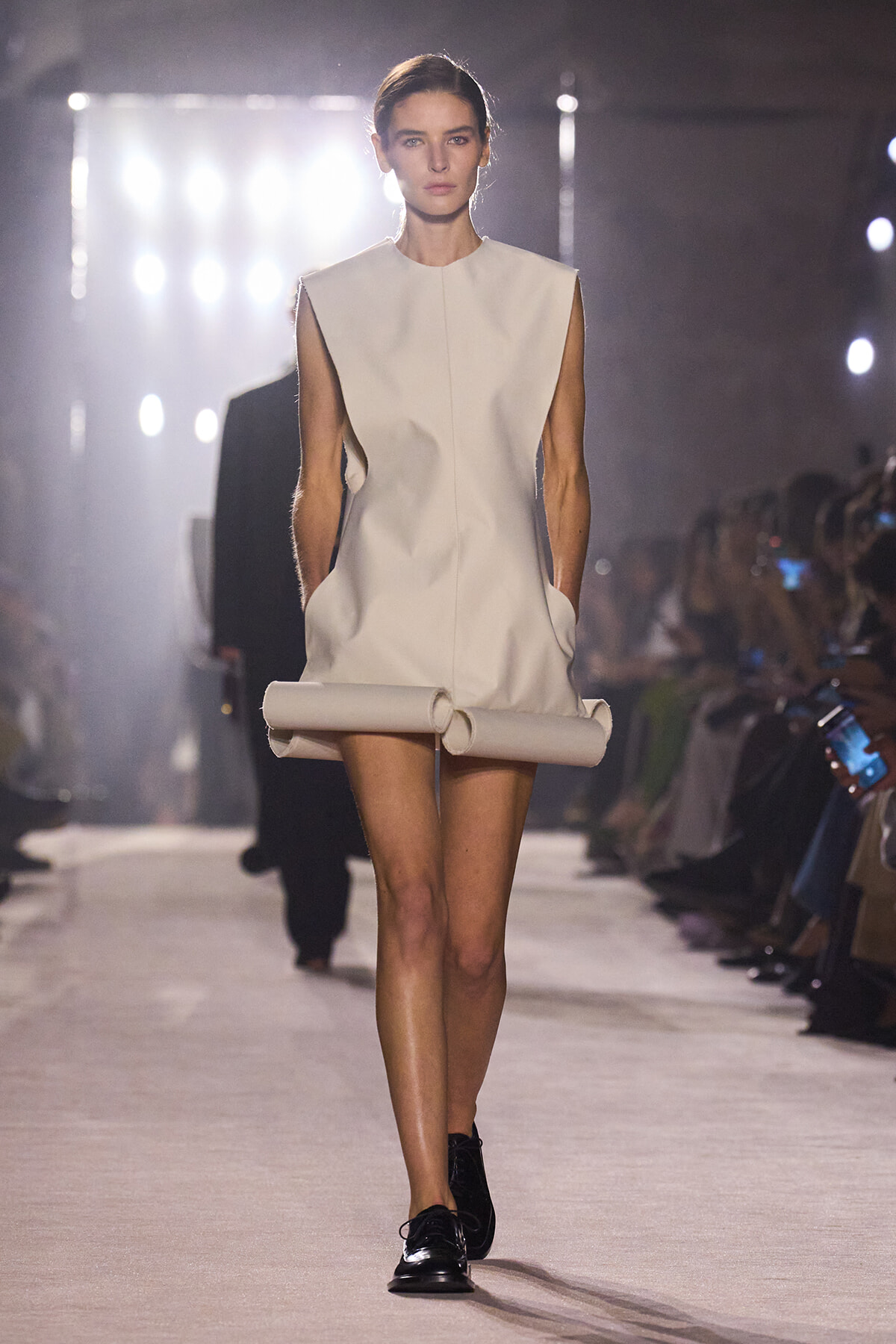 Model walks a runway in a minimalist sleeveless ivory shift dress with a high, round neckline and sculpted shoulders, paired with black lace-up shoes and a rolled fabric accessory at the hem.