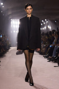 Model on a runway wearing a dark oversized blazer over a black outfit, sheer tights, black heels, and a small gray clutch.