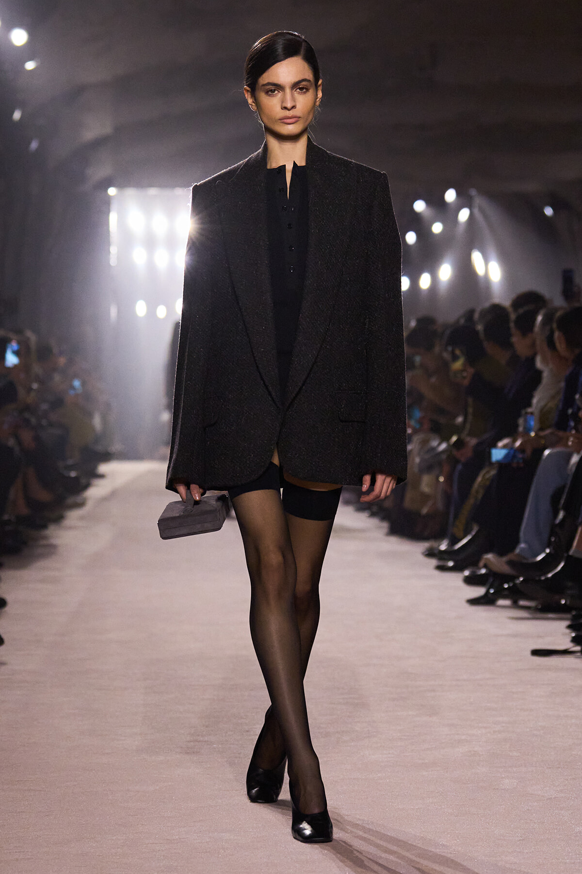 Model on a runway wearing a dark oversized blazer over a black outfit, sheer tights, black heels, and a small gray clutch.