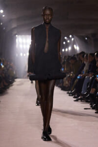 Black sleeveless mini dress with a large rolled hem and teardrop keyhole neckline, worn by a model walking a runway.