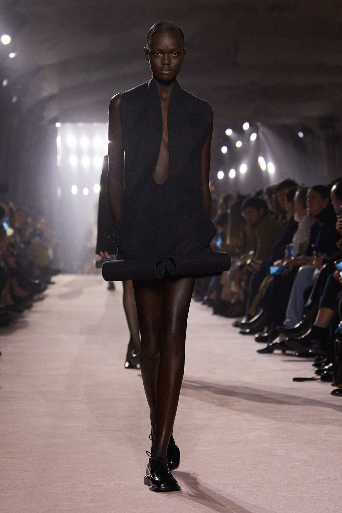 Black sleeveless mini dress with a large rolled hem and teardrop keyhole neckline, worn by a model walking a runway.