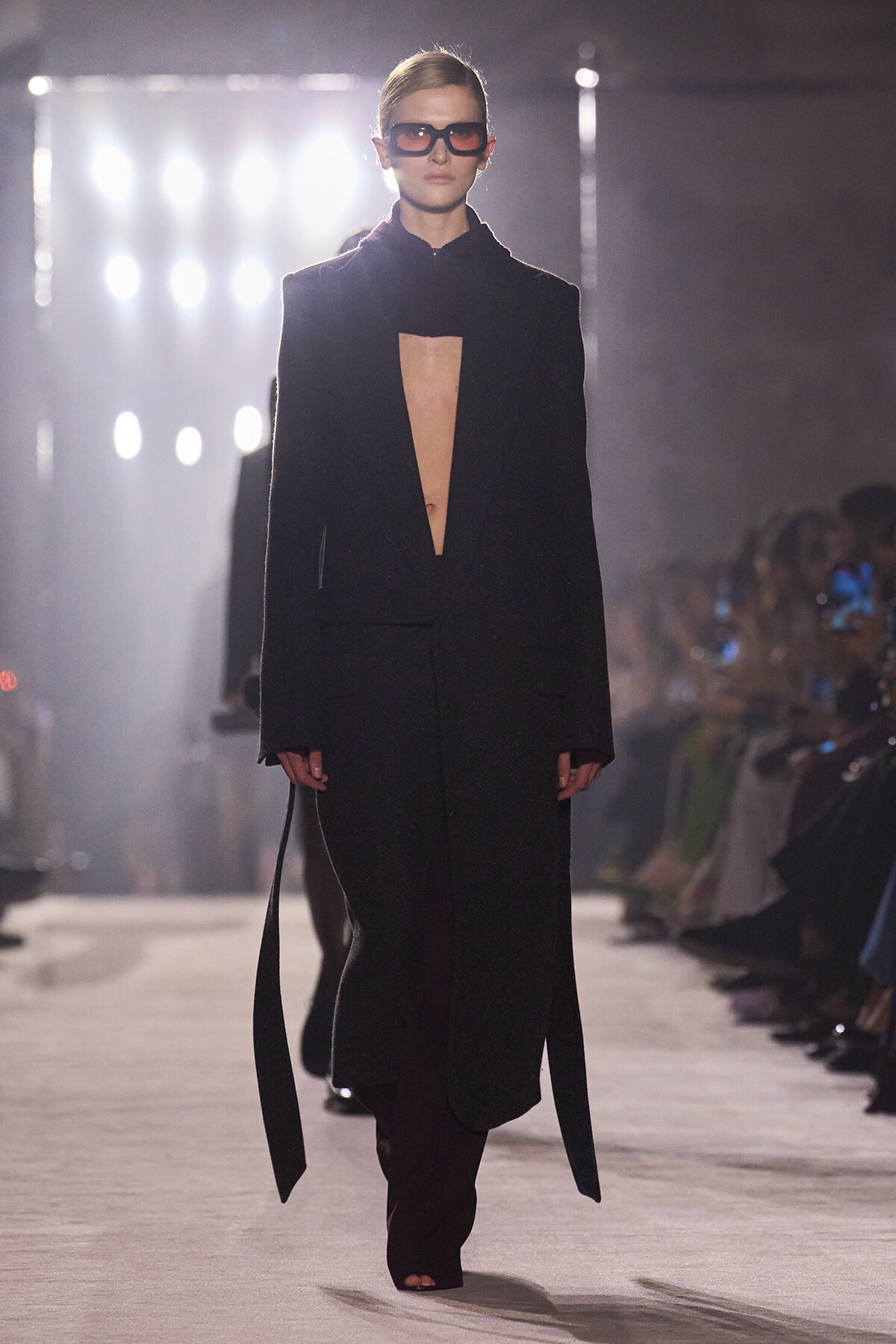 Runway model in a black long coat with a deep front opening, paired with dark sunglasses and a sleek updo.