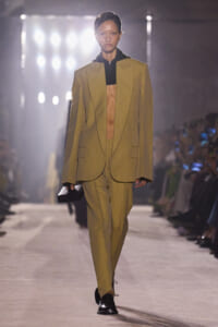Model walks the runway in a loose mustard suit with an open blazer over a black cropped top, carrying a small black clutch.