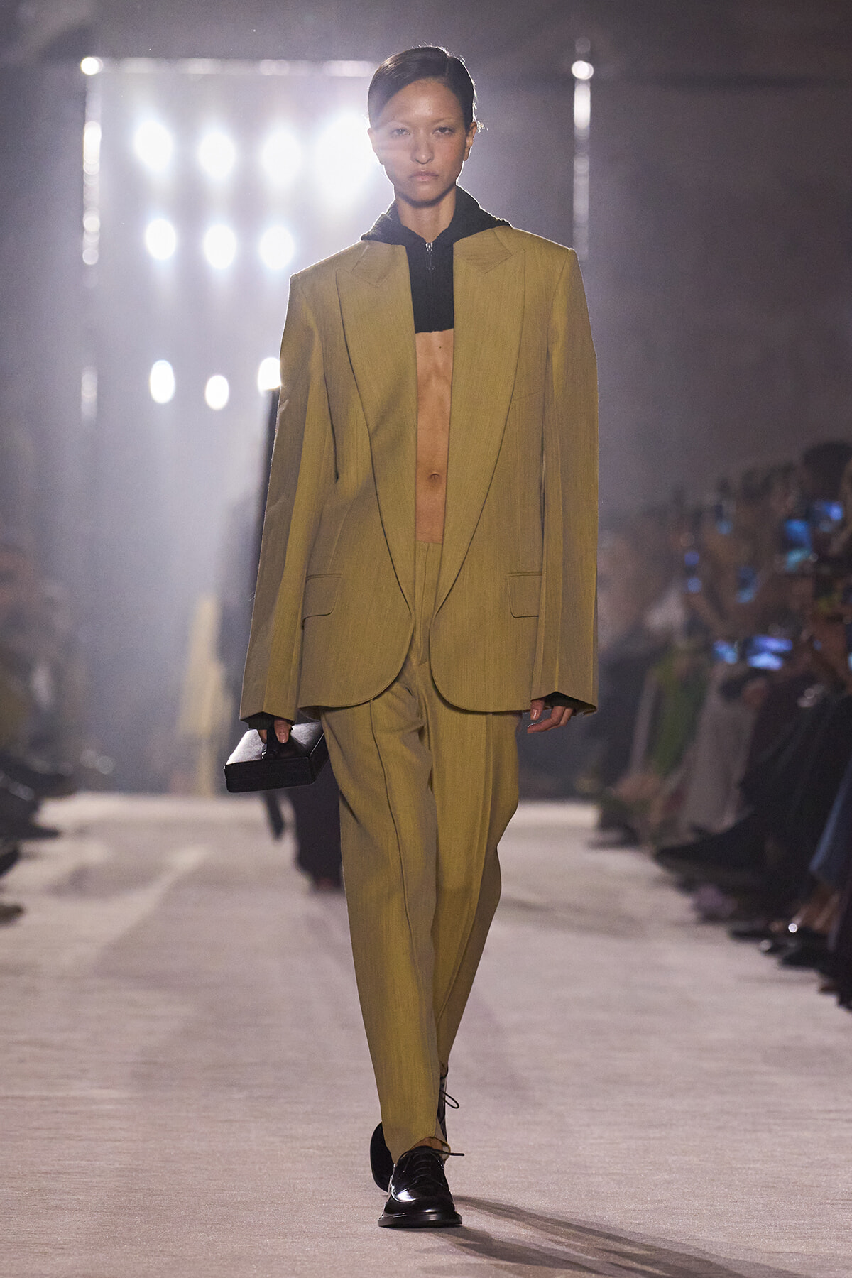 Model walks the runway in a loose mustard suit with an open blazer over a black cropped top, carrying a small black clutch.