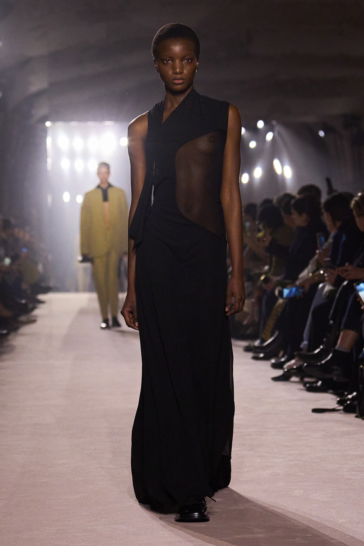 Model walks a fashion show in a black sheer dress with cutouts on a dimly lit runway.