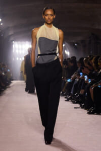 Model on a runway wearing a beige sheer halter top over a gray panel bodice with black high-waisted trousers, hands in pockets while spectators watch.