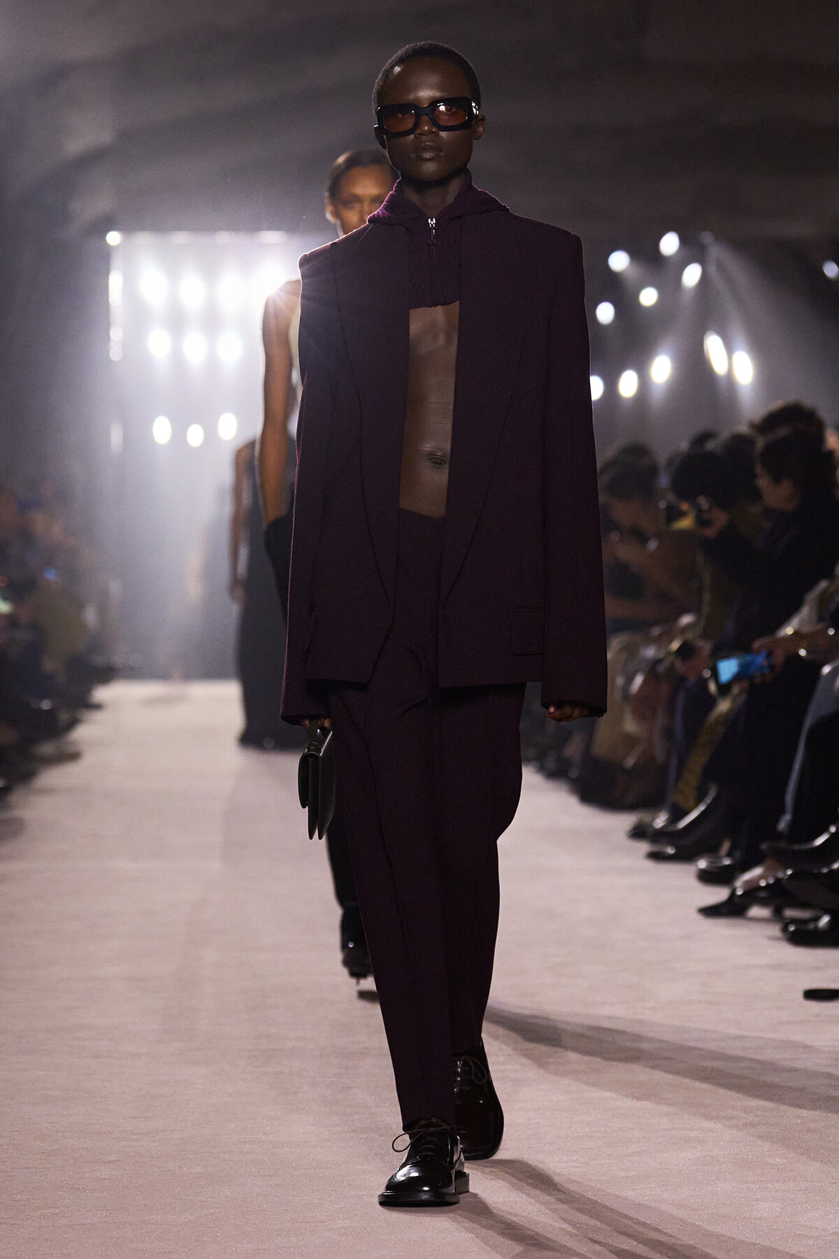 Model walking a runway in a dark burgundy pantsuit with an oversized cape, black sunglasses, and a small black handbag, backstage lighting in the background.