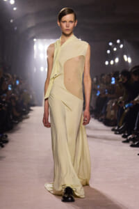 Model walks the runway in a pale yellow draped gown with a sheer side panel and dark shoes against a bright fashion-show backdrop.