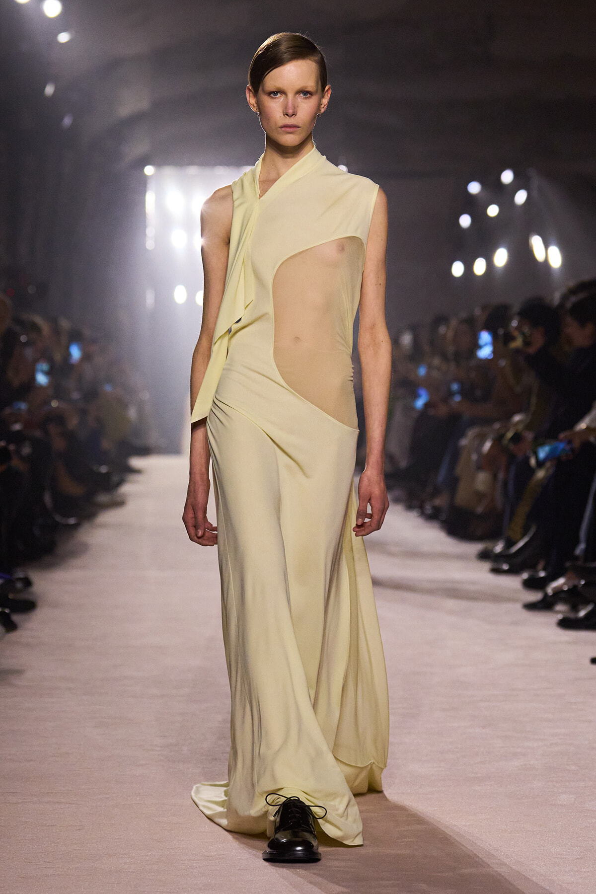 Model walks the runway in a pale yellow draped gown with a sheer side panel and dark shoes against a bright fashion-show backdrop.