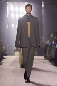 Model walks a runway in an oversized gray suit with a long blazer, open at the chest, while holding a small rectangular clutch.
