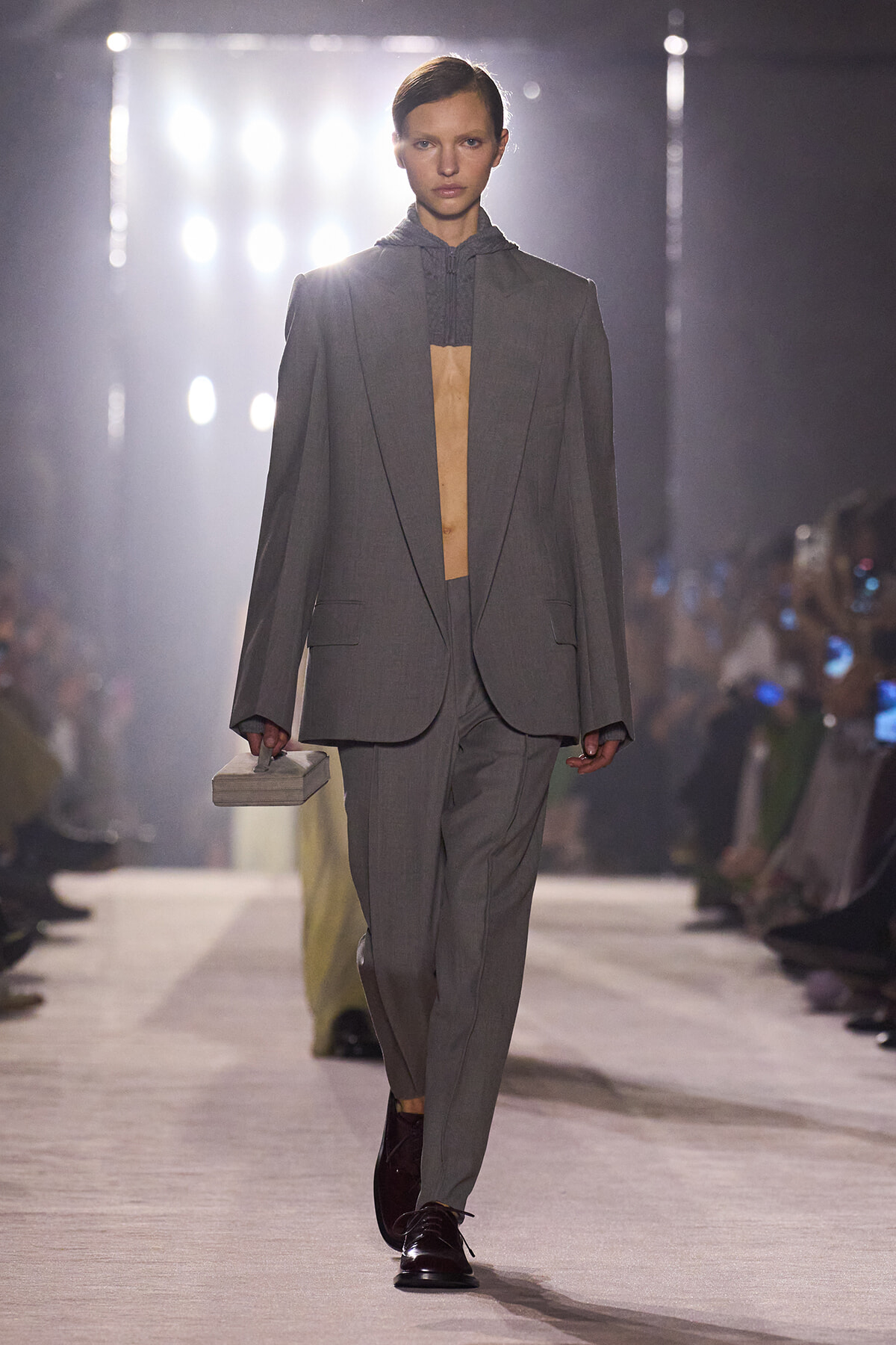 Model walks a runway in an oversized gray suit with a long blazer, open at the chest, while holding a small rectangular clutch.