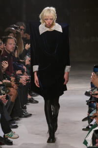 Model walking a fashion runway in a dark velvet dress with oversized white lace collar and cuffs, surrounded by seated spectators and photographers.