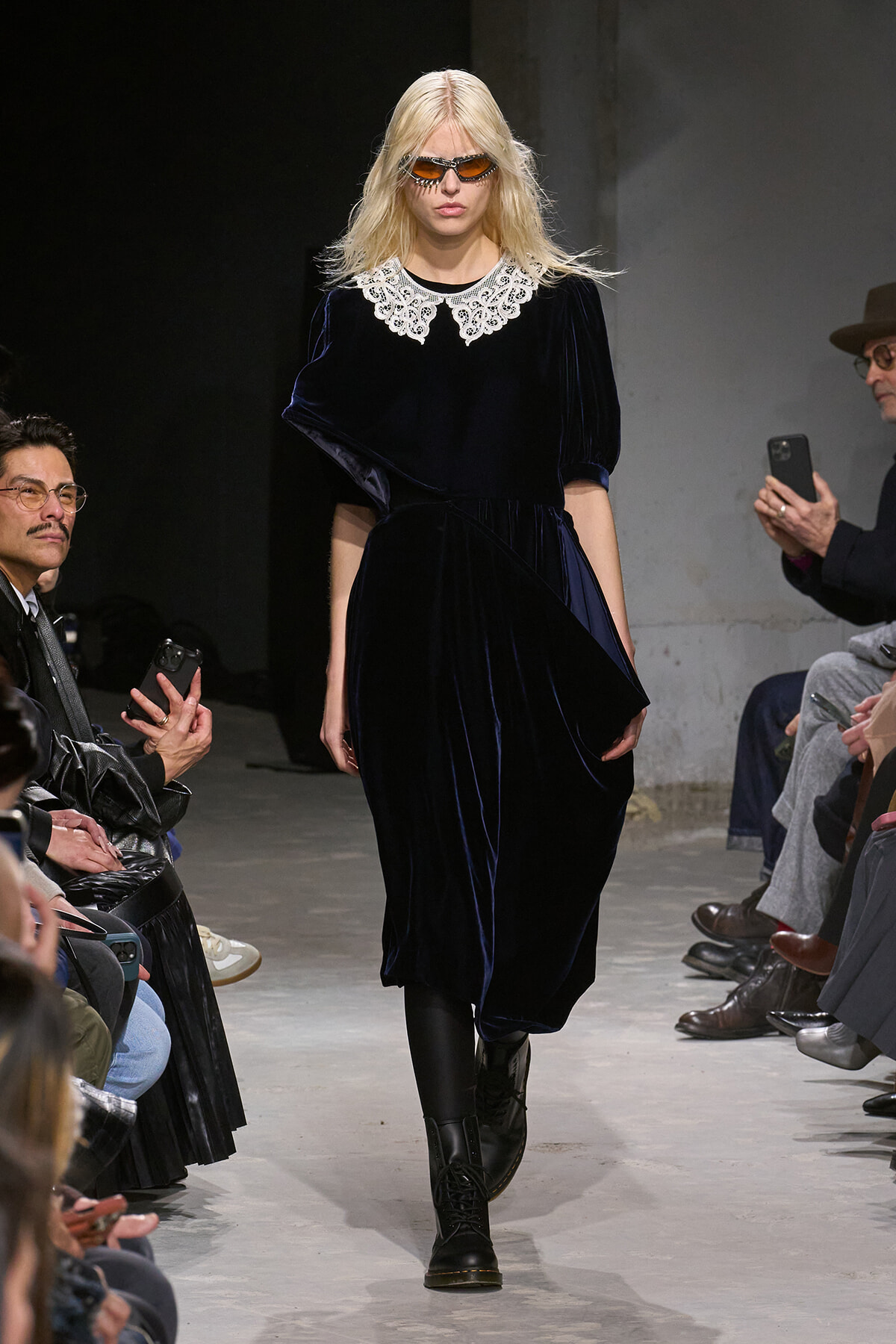 Model on a runway wearing a navy velvet dress with a white lace collar, black boots, and orange sunglasses.