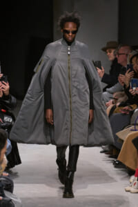 Model walks the runway in a large gray cape-style coat with a front zipper, over a black outfit and sunglasses.