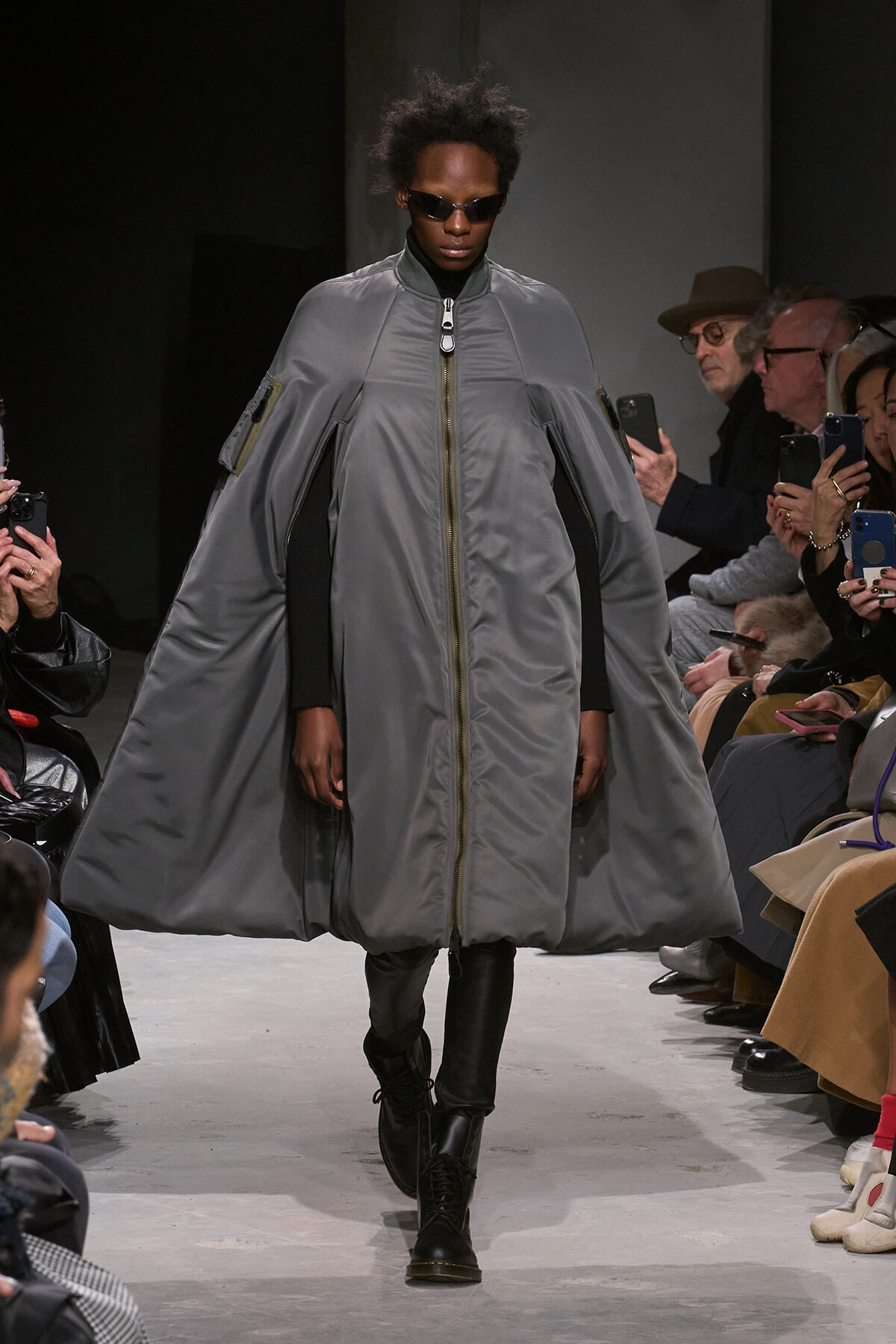 Model walks the runway in a large gray cape-style coat with a front zipper, over a black outfit and sunglasses.