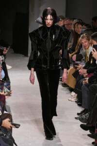 Model walks a runway in a glossy black geometric jacket with oversized, angular shoulders and matching black pants.
