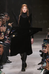 Model walks the runway in a long black velvet dress with angular black panels and dramatic sculptural accessories, audience fans taking photos on both sides.