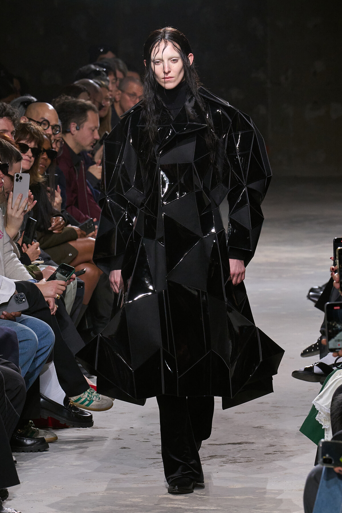 Model on a runway wearing a shiny black, faceted coat made of angular panels, with spectators along the sides of the catwalk.
