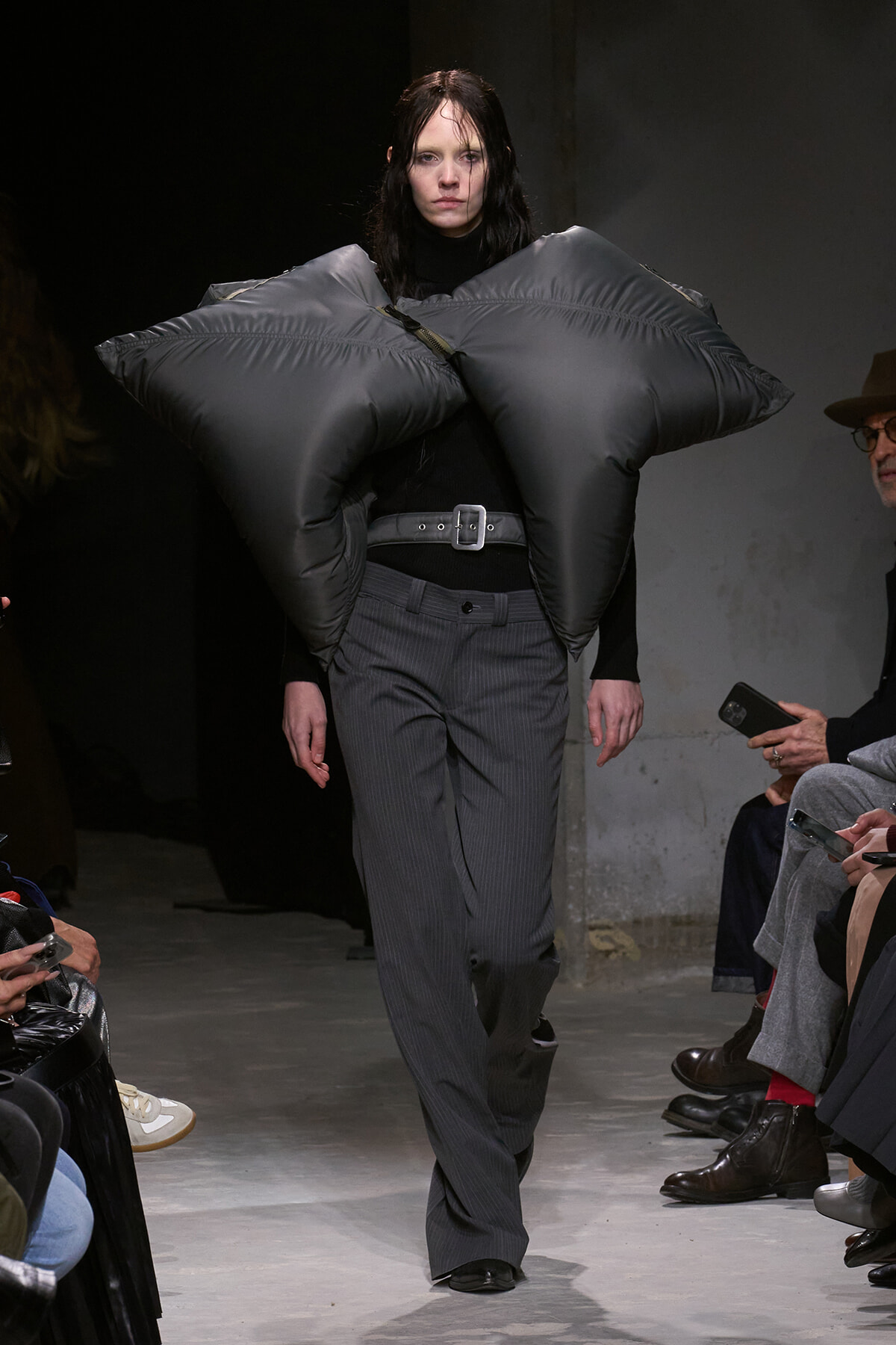 Model on a runway wearing a dramatic oversized gray padded shoulder piece over a black turtleneck and pinstripe trousers. Audience visible along the sides.
