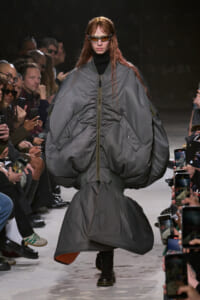 Model walks runway in oversized gray puffer coat with zipper, sunglasses, and black turtleneck, audience filming nearby