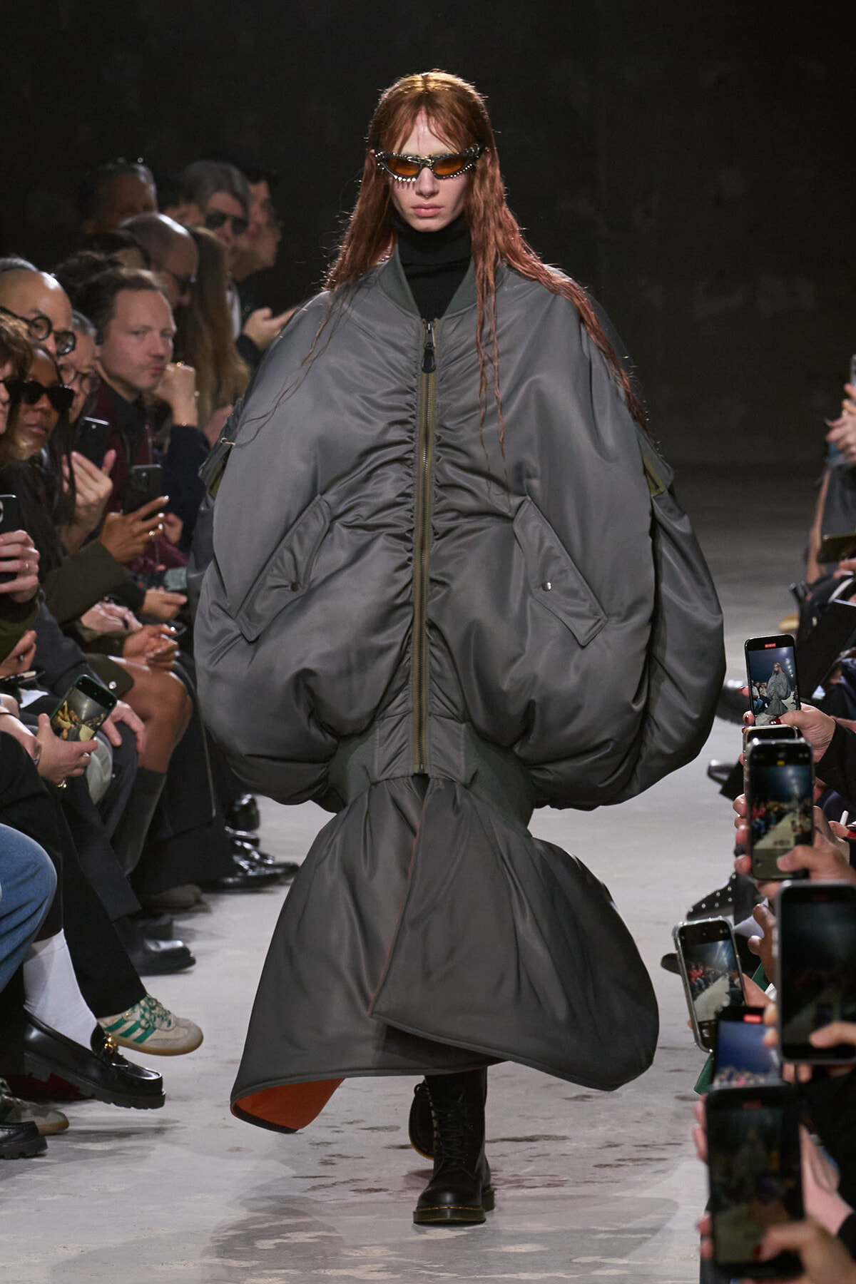 Model walks runway in oversized gray puffer coat with zipper, sunglasses, and black turtleneck, audience filming nearby