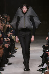 Runway model wearing an oversized gray padded vest with high, wing-like shoulders over a black turtleneck and pants.