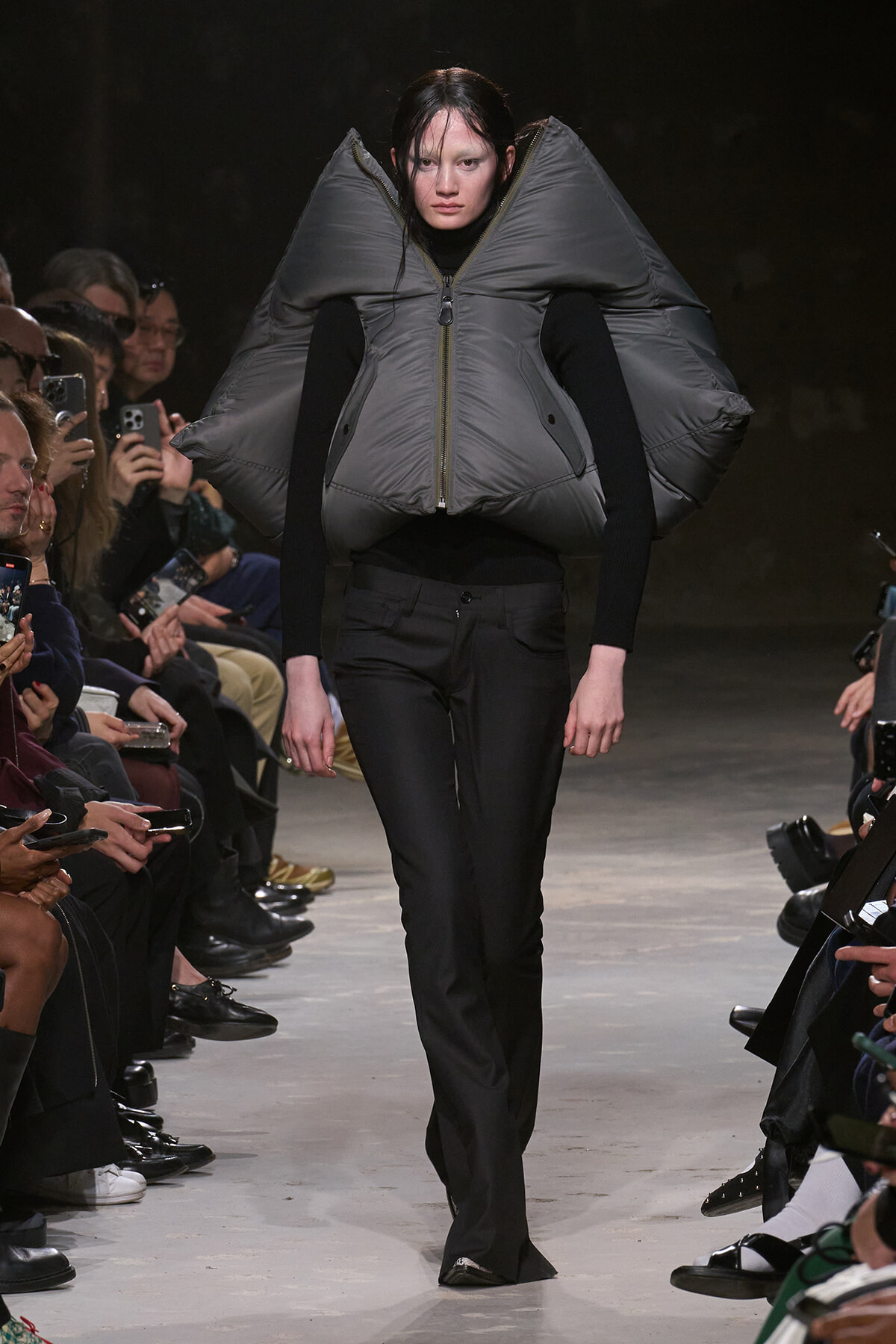 Runway model wearing an oversized gray padded vest with high, wing-like shoulders over a black turtleneck and pants.