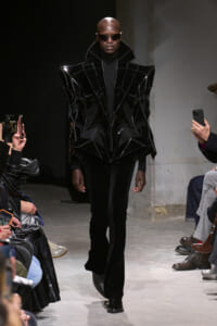 Model on runway wearing a glossy black geometric cape-like jacket with high collar and sunglasses, paired with black pants and boots, in a dark industrial setting.