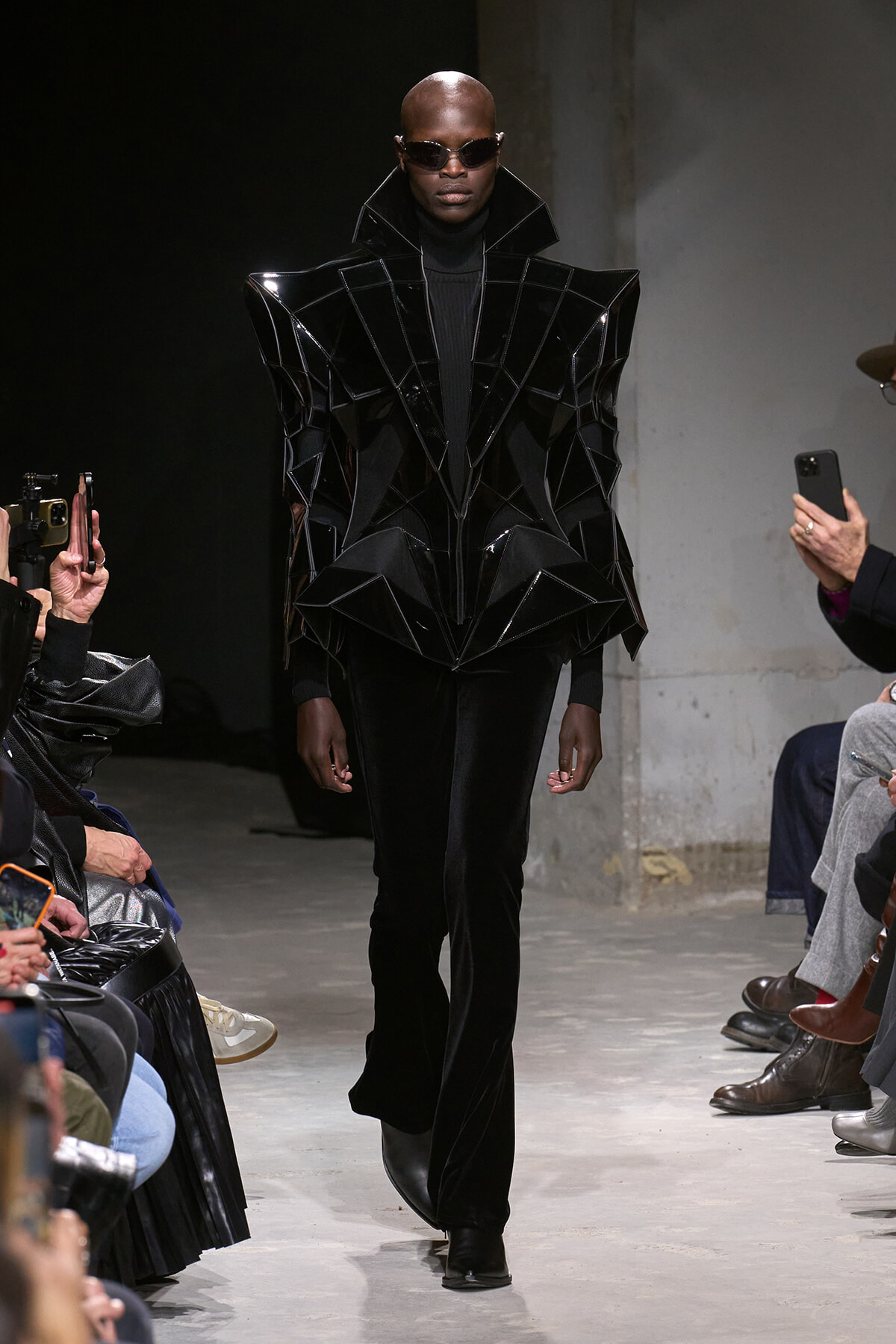 Model on runway wearing a glossy black geometric cape-like jacket with high collar and sunglasses, paired with black pants and boots, in a dark industrial setting.