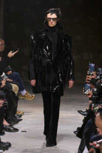 Model walking the runway in a glossy, geometric black coat with a black turtleneck and sunglasses; audience members hold up phones to record.