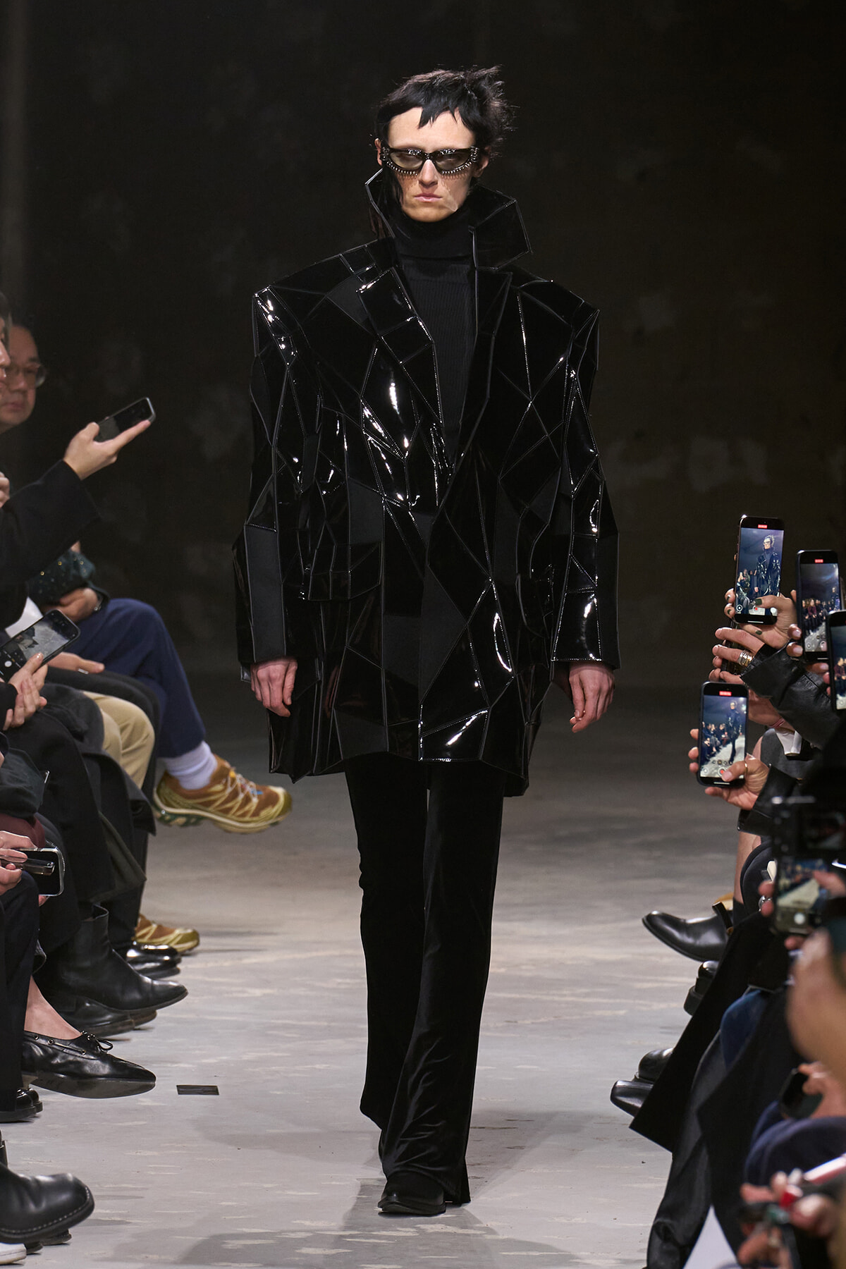 Model walking the runway in a glossy, geometric black coat with a black turtleneck and sunglasses; audience members hold up phones to record.