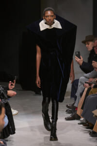 Runway model in a navy velvet dress with an oversized white lace collar, pearl necklace, and black boots, walking the catwalk.