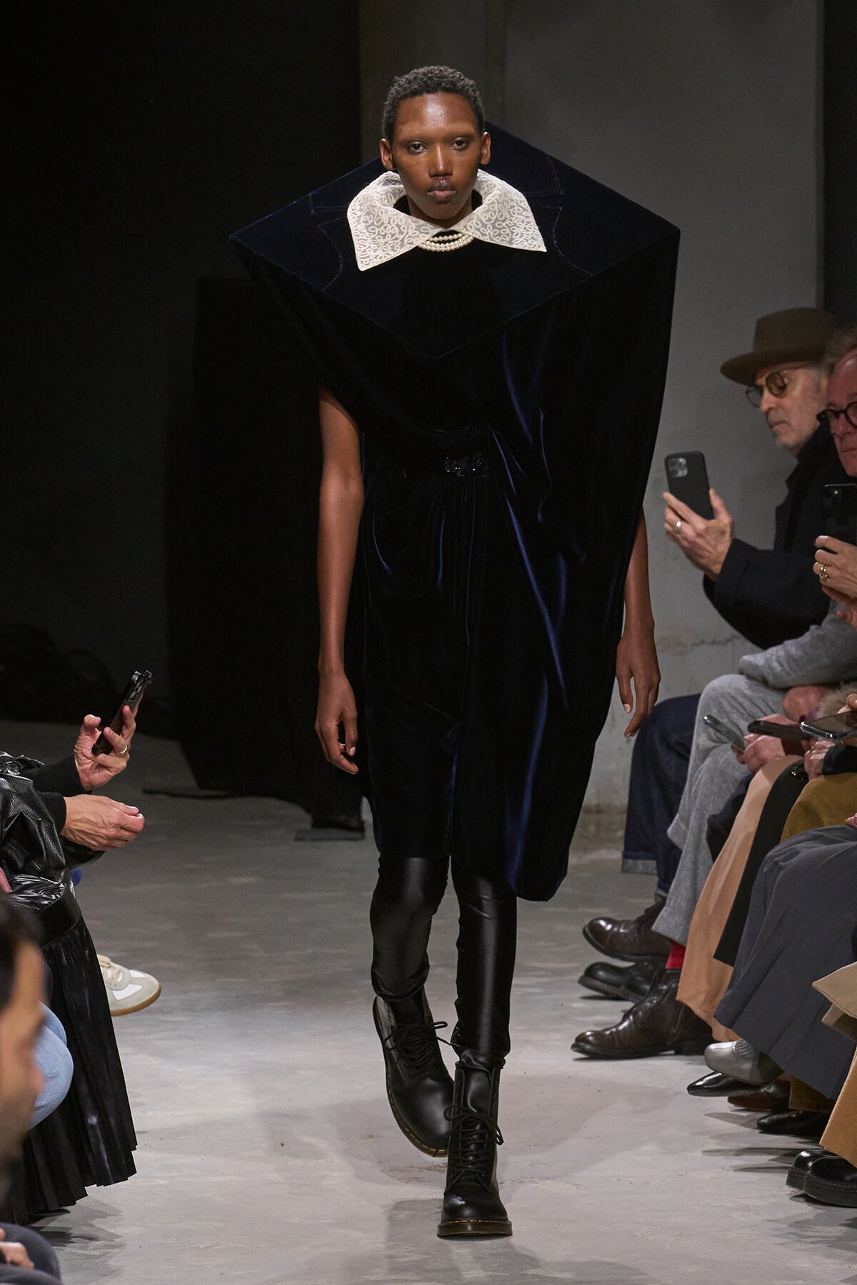 Runway model in a navy velvet dress with an oversized white lace collar, pearl necklace, and black boots, walking the catwalk.