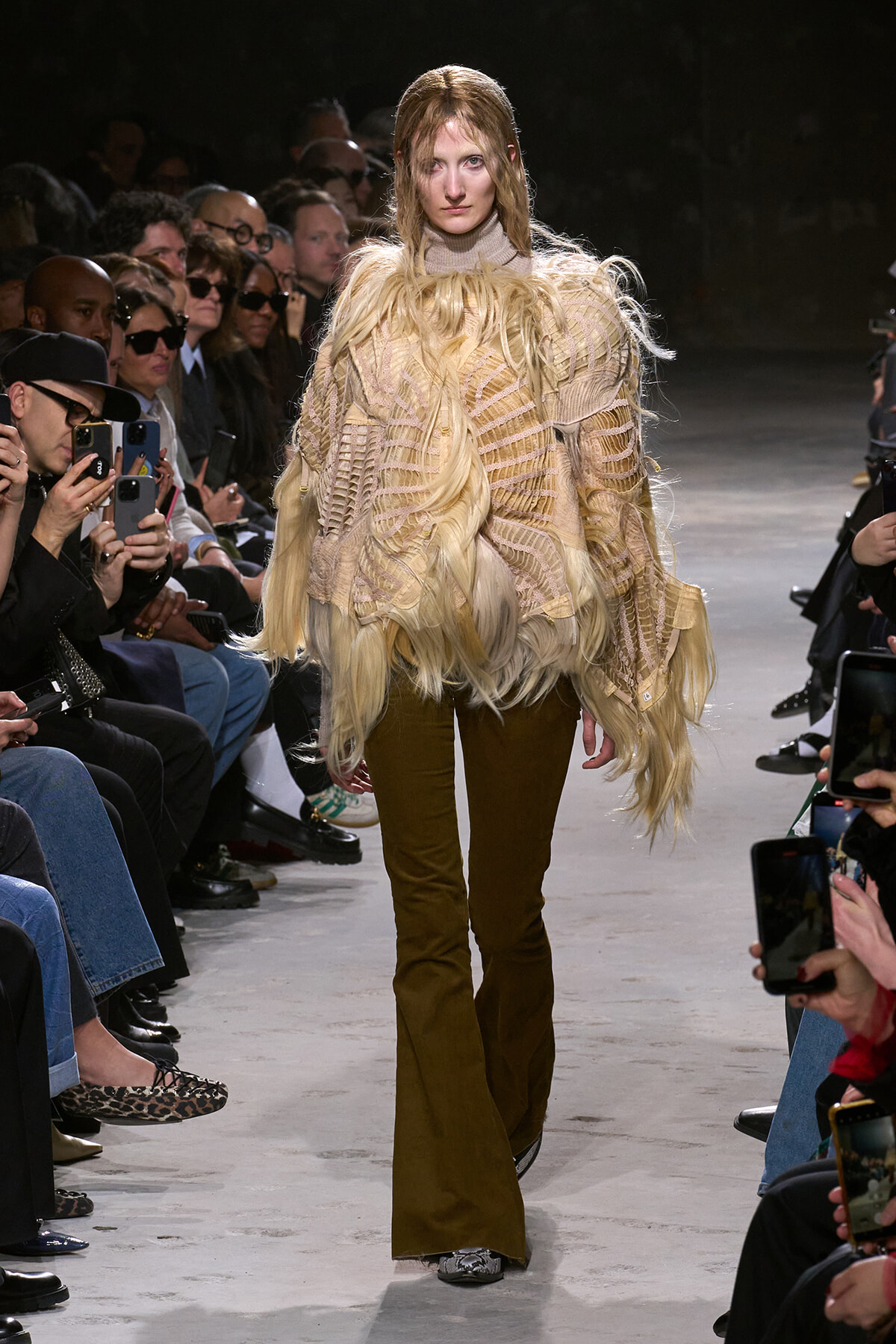Model walking a fashion runway in a beige fringed knit top and brown flared pants, with long blonde hair, surrounded by an audience taking photos.