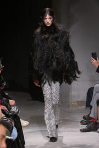 Model on runway wearing a black feathered oversized top and snakeskin-print wide-leg pants on the catwalk with photographers nearby.