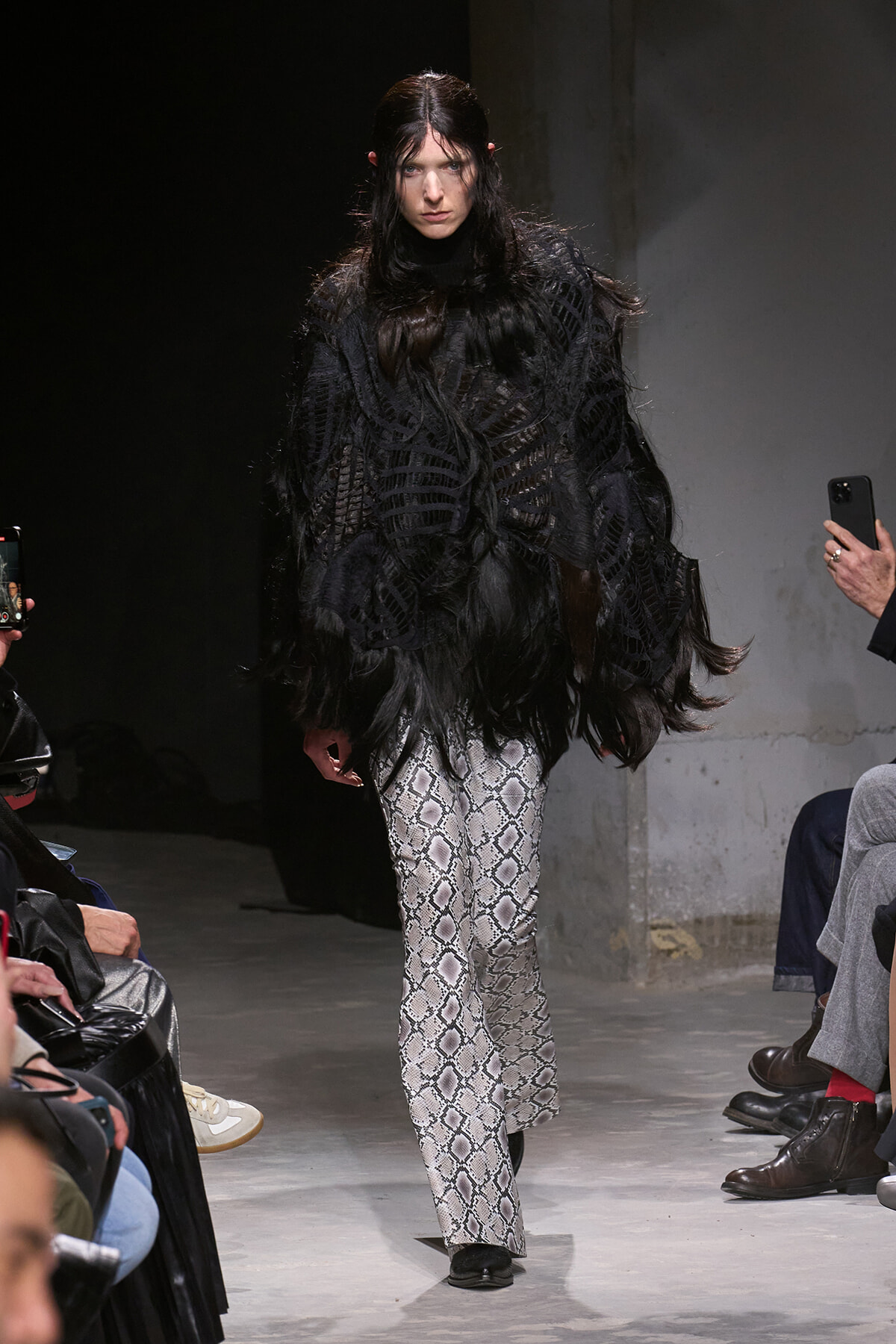 Model on runway wearing a black feathered oversized top and snakeskin-print wide-leg pants on the catwalk with photographers nearby.