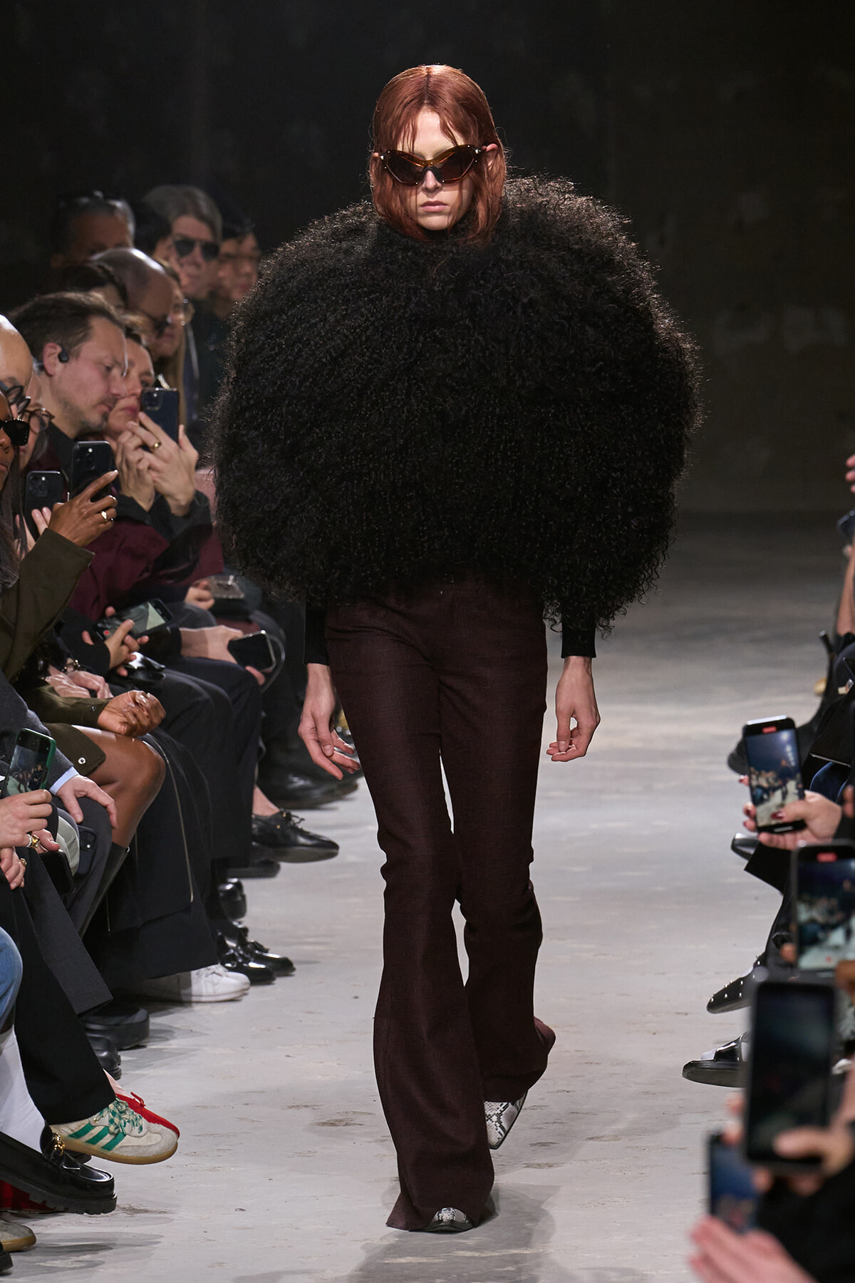 Model walking a fashion show runway in a voluminous black furry top and burgundy flared pants, wearing oversized sunglasses; audience lines the catwalk filming.