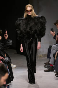 Model on a runway wearing a dramatic black feathered coat over a black outfit with velvet pants and oversized brown sunglasses.