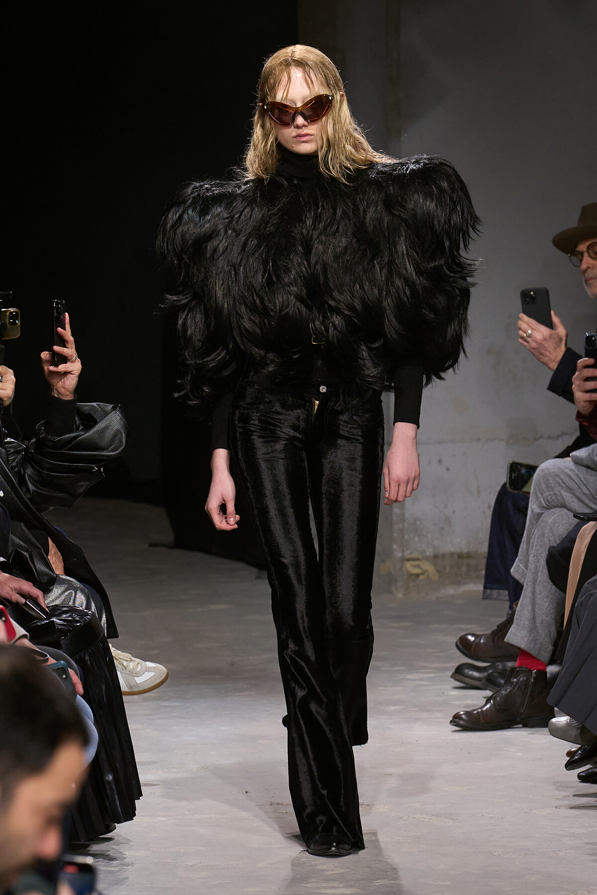 Model on a runway wearing a dramatic black feathered coat over a black outfit with velvet pants and oversized brown sunglasses.