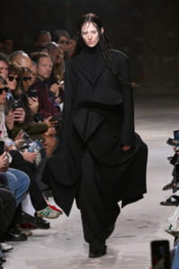 Model walks a runway in an oversized black sculptural coat as the audience watches and photographs her.